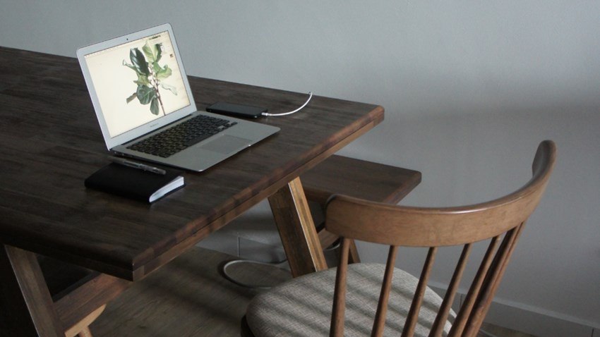 Scrivania in legno con un portatile, un taccuino e una sedia in un ambiente di lavoro semplice.